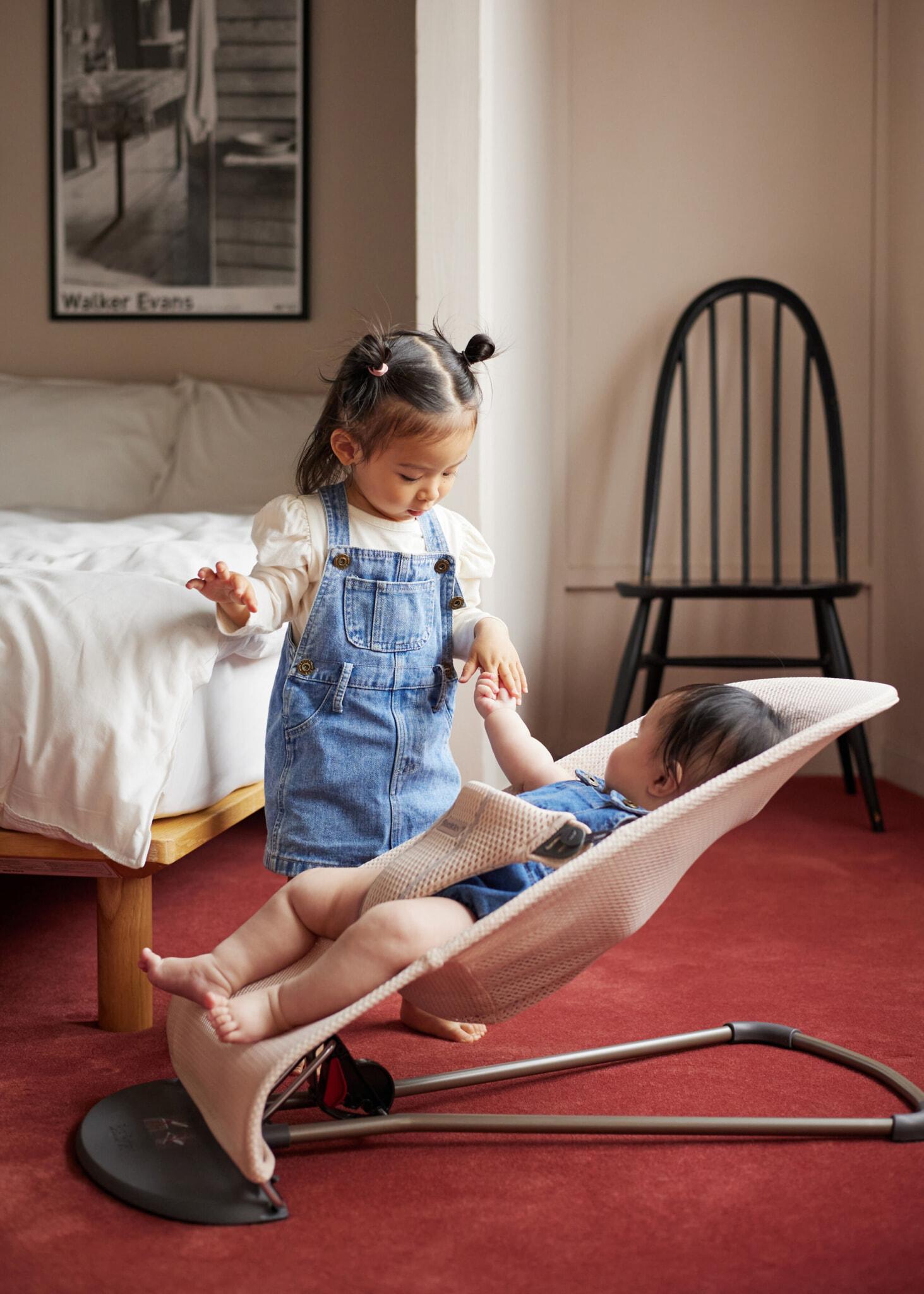 A toddler in denim overalls gently touches a baby in a bouncer. They are in a cozy room with a bed and a chair in the background.