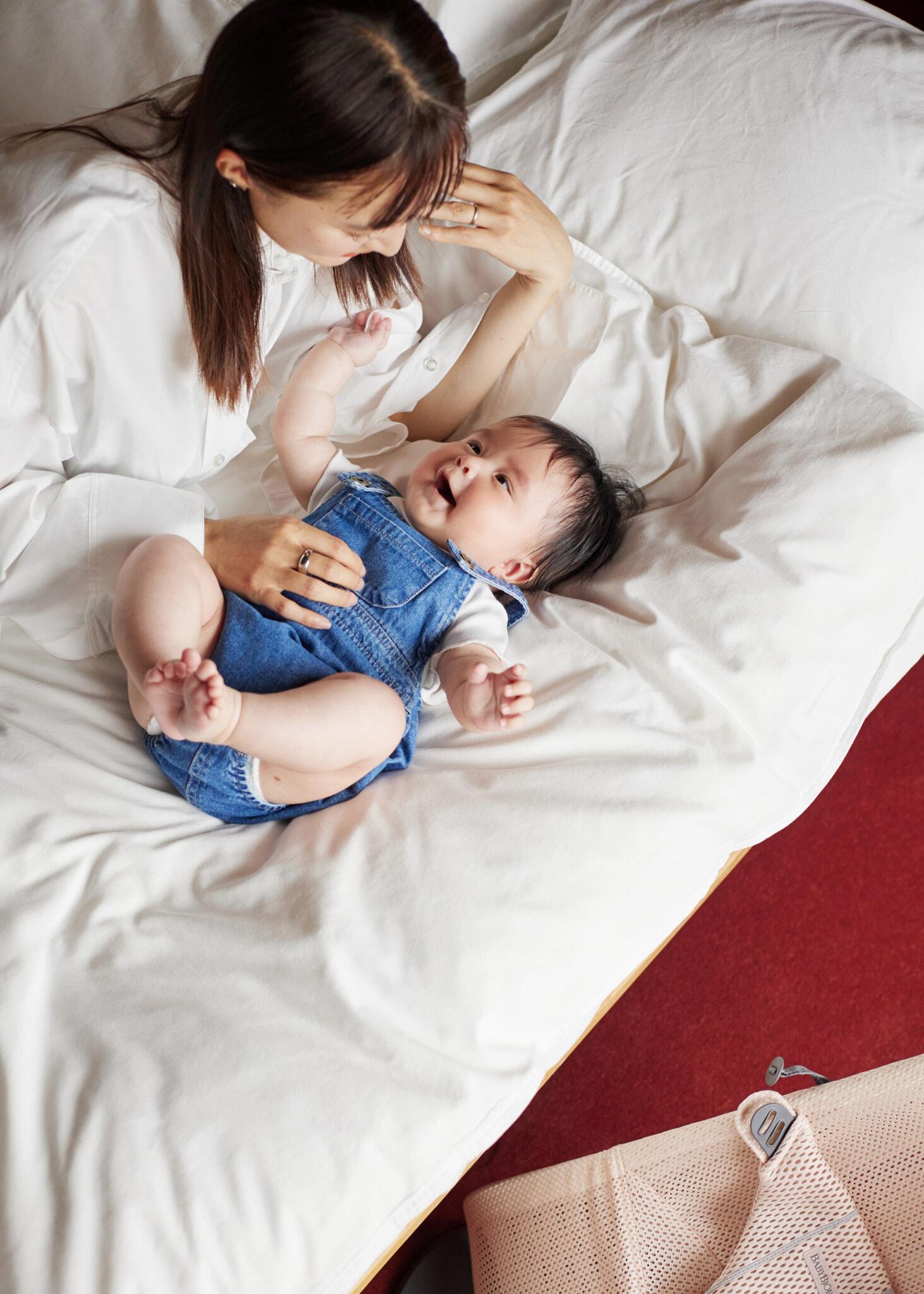 A woman in a white shirt lovingly gazes at a baby in denim overalls, lying on a white bed with a red floor visible.