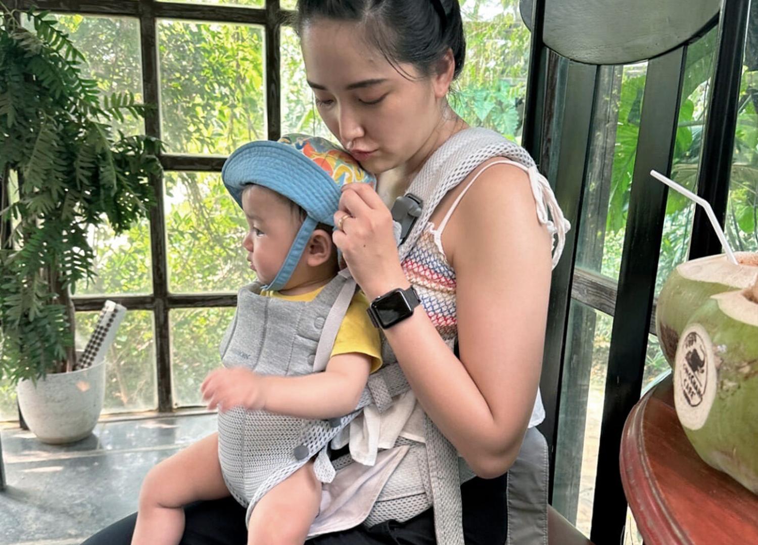 Woman sitting in a sunlit room, adjusting a blue hat on a baby in a carrier. They are near a table with a coconut drink.