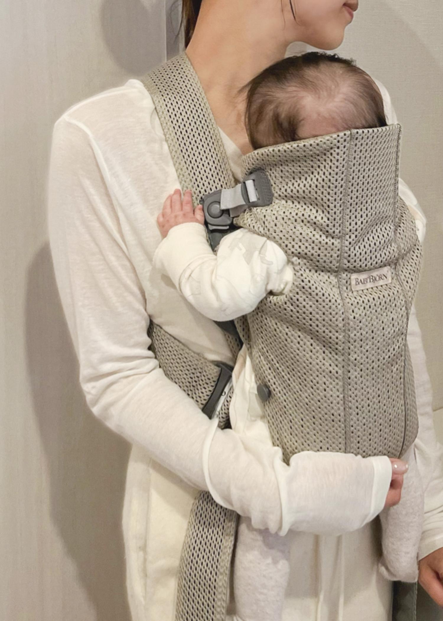 Woman wearing a white shirt holds a baby in a gray baby carrier, facing forward.