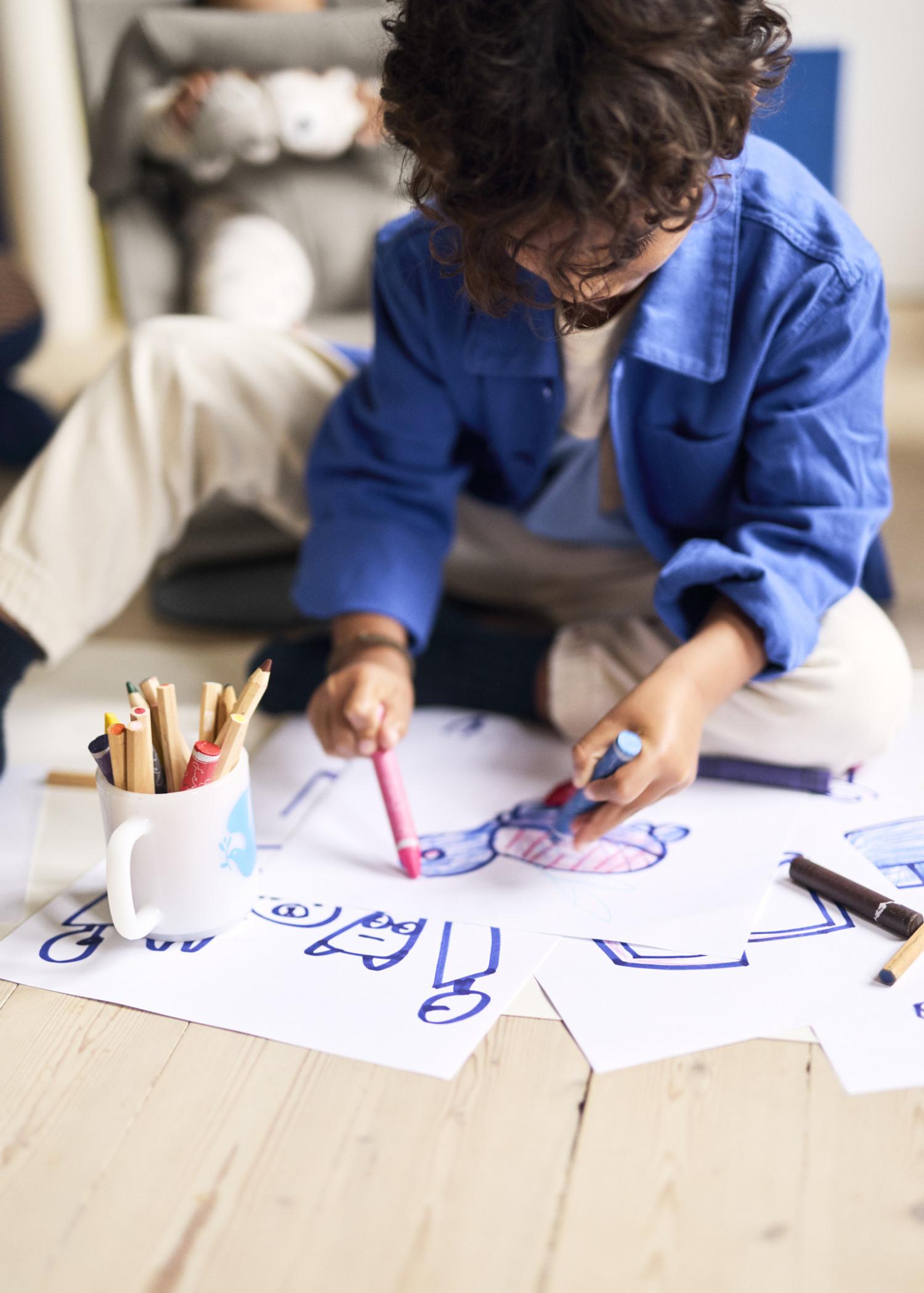 Child drawing with markers on paper, surrounded by colored pencils in a cup, sitting on a wooden floor.