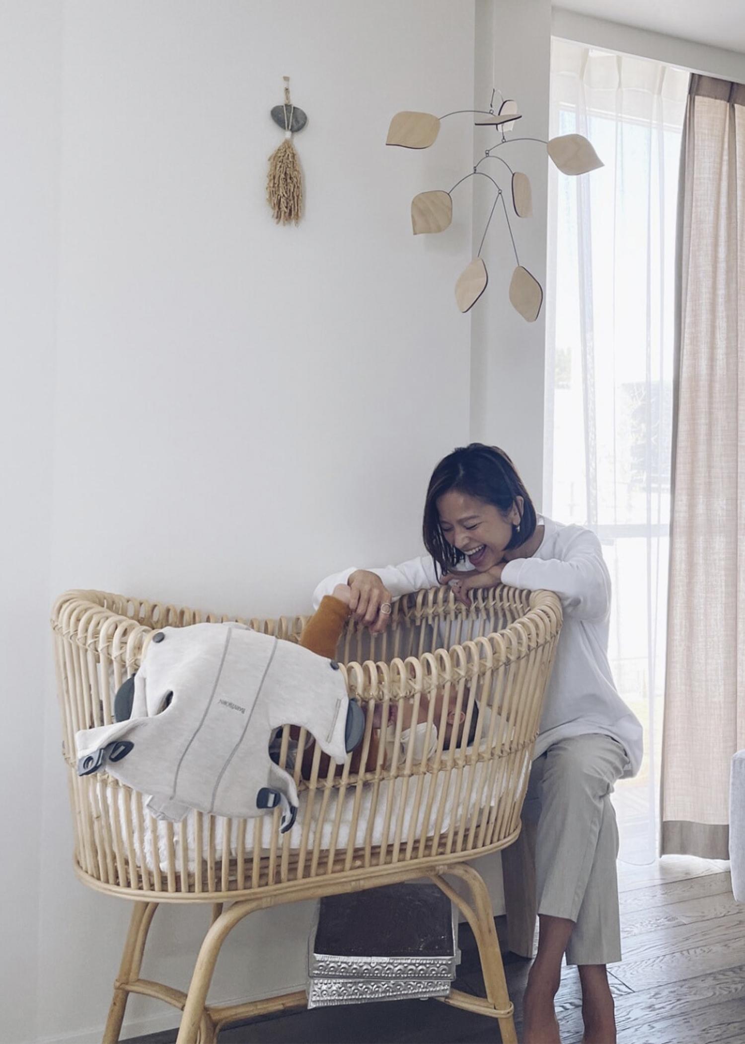 A woman smiles at a baby in a wicker bassinet, in a bright room with wooden floors and sheer curtains. A mobile hangs above the bassinet.