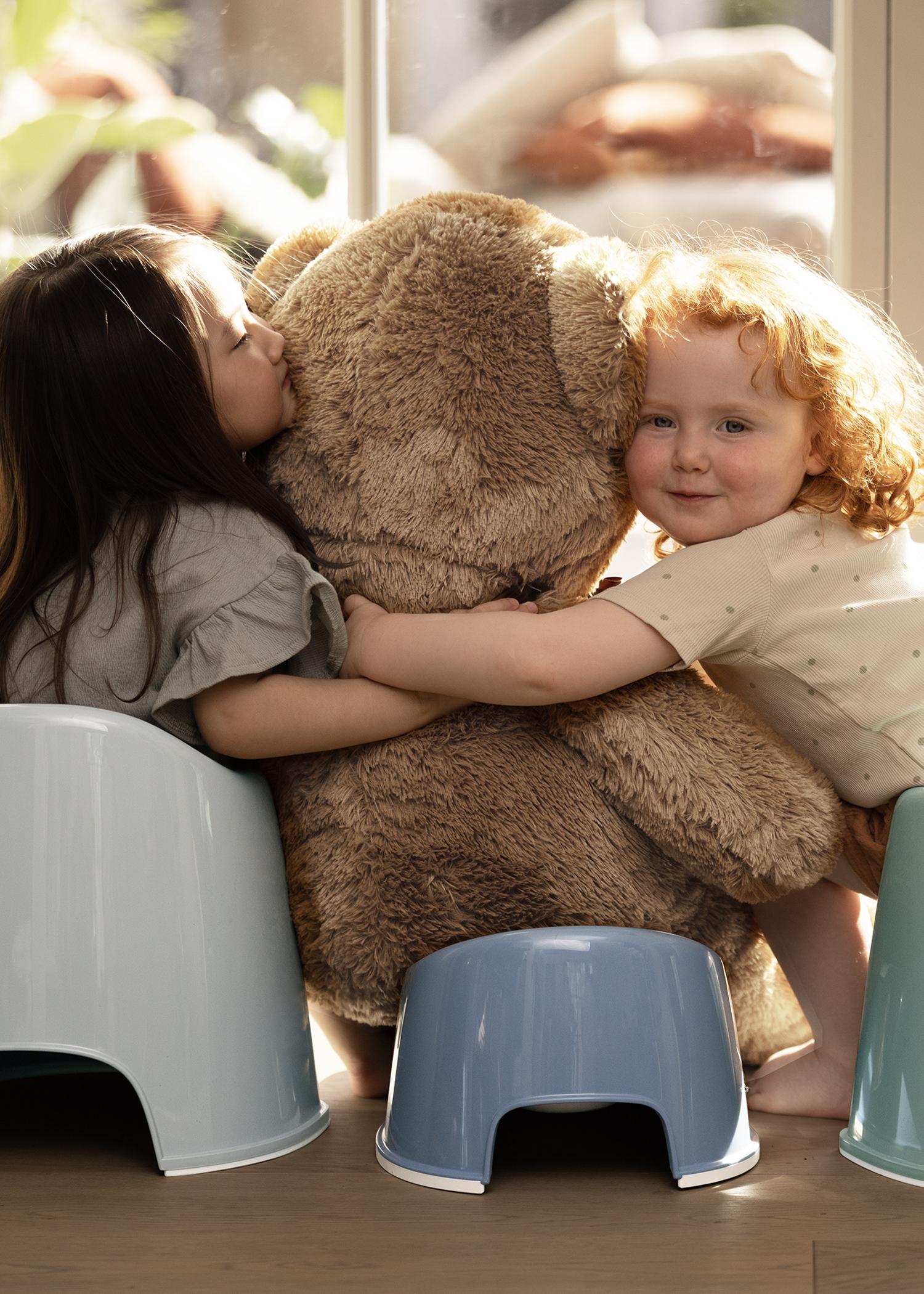Two children hugging a large teddy bear while sitting on small stools, with sunlight streaming through a window in the background.
