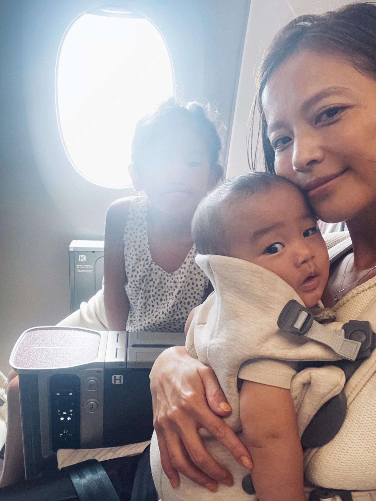 A woman holds a baby, seated next to a child by an airplane window. They appear relaxed and content during the flight.