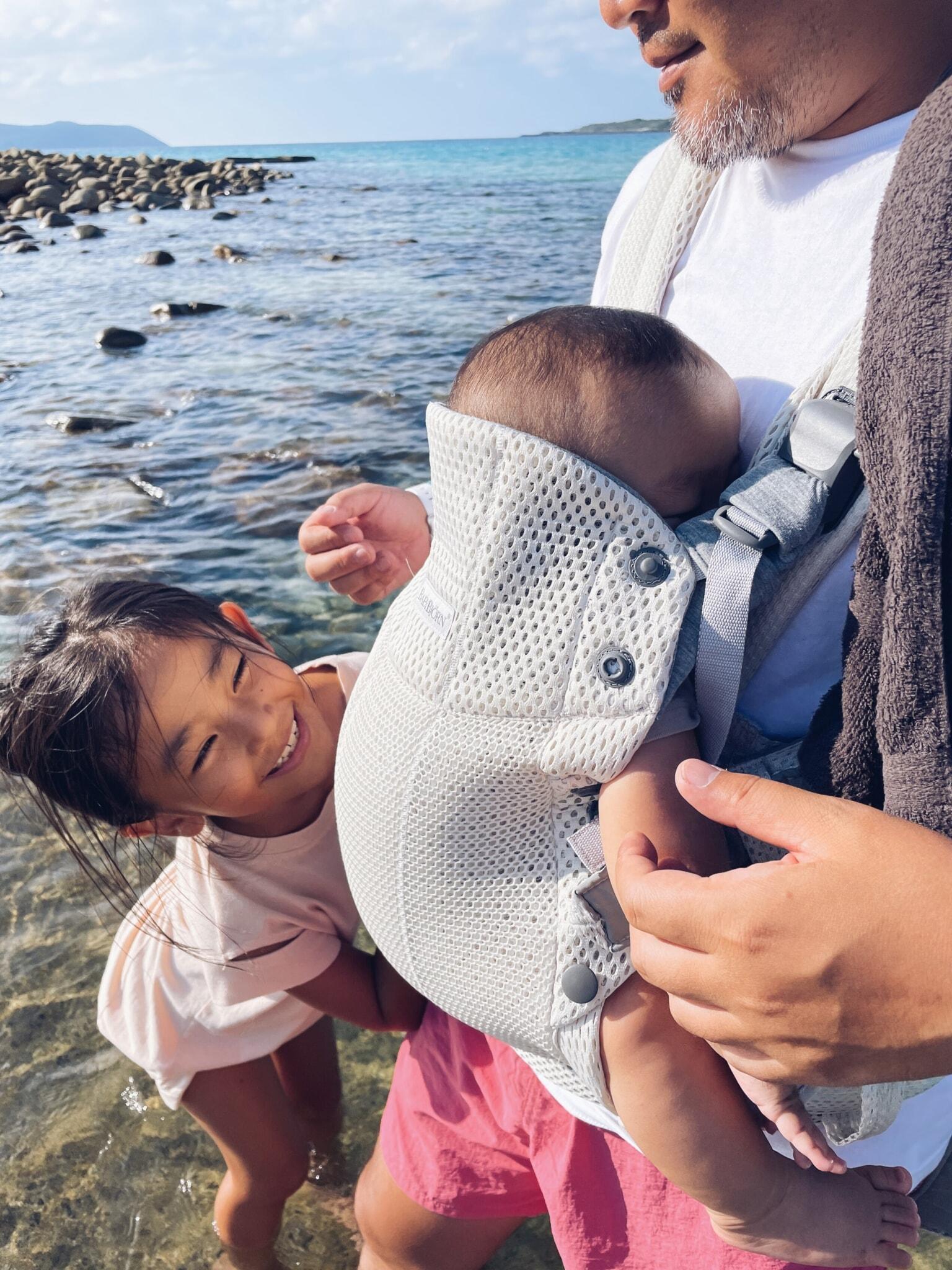 A man stands by the water holding a baby in a carrier. A smiling girl leans in, looking at the baby. Rocky shoreline and blue sea in the background.
