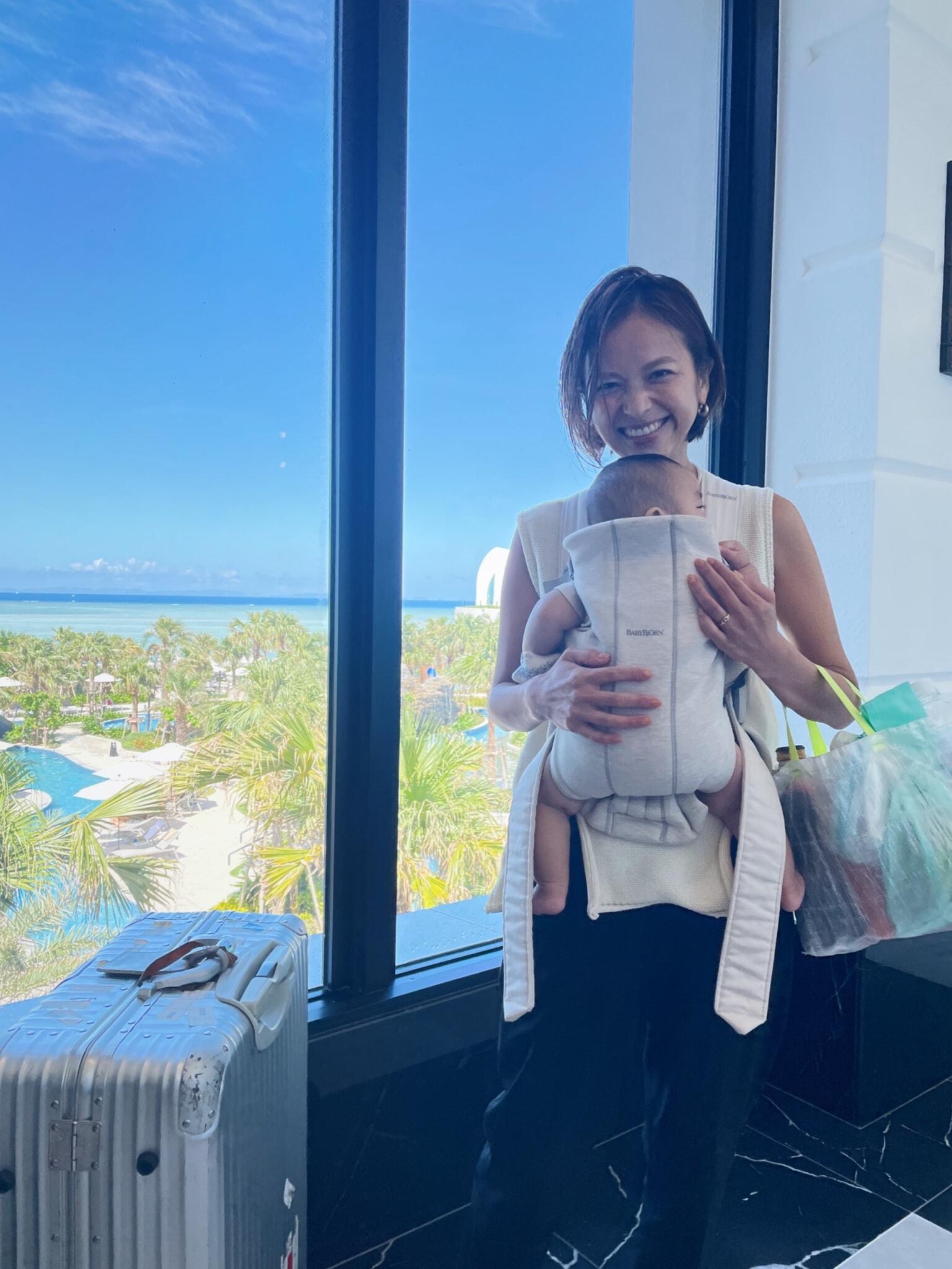 A woman smiling while holding a baby in a carrier, standing by a window with a view of a pool and palm trees. A suitcase is beside her.