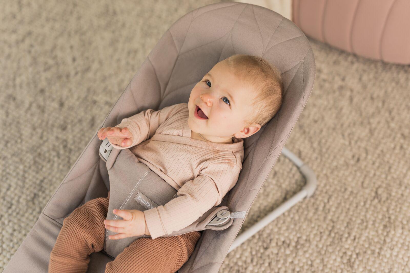 A smiling baby in a beige bouncer, wearing a light pink top and brown pants, sits on a textured carpet.