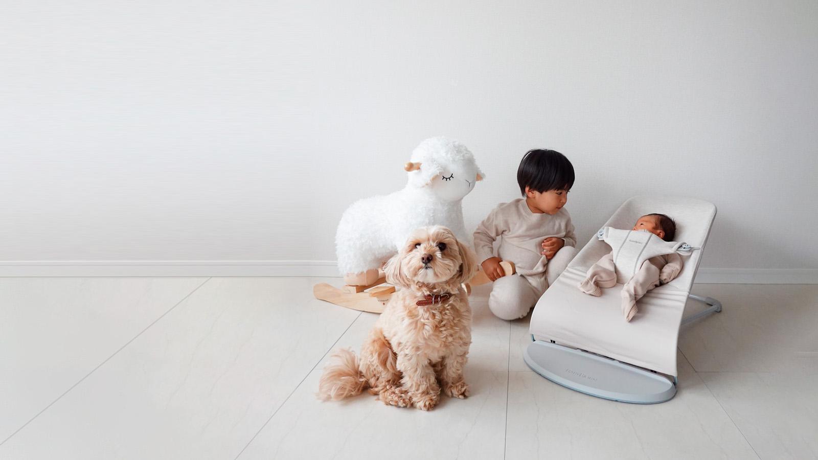 A child sits beside a baby in a rocker, with a fluffy dog and a large stuffed sheep nearby on a light-colored floor.