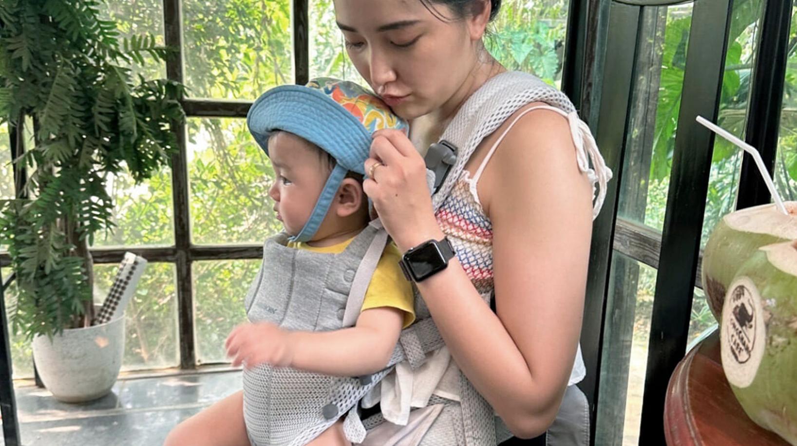 A woman sits holding a baby in a carrier, adjusting the baby's blue cap. They are in a sunlit room with plants and a coconut drink nearby.