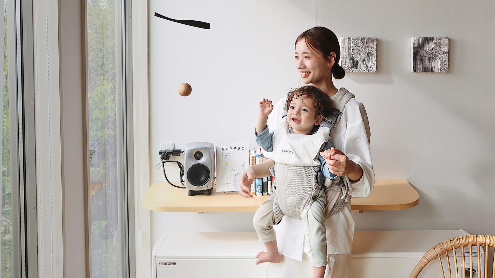 A smiling woman stands by a window, holding a happy toddler in a baby carrier. The child reaches for a ball in mid-air.