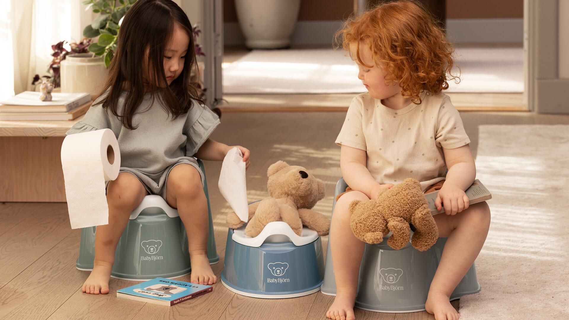 Two children sitting on potties, each holding a teddy bear. One has a toilet paper roll, and a book is on the floor.