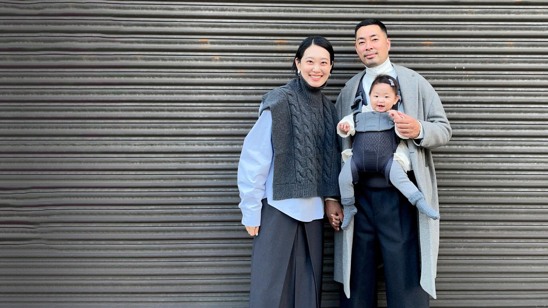 A smiling couple stands in front of a corrugated metal wall, holding a baby in a carrier. They are dressed in coordinated gray and black outfits.