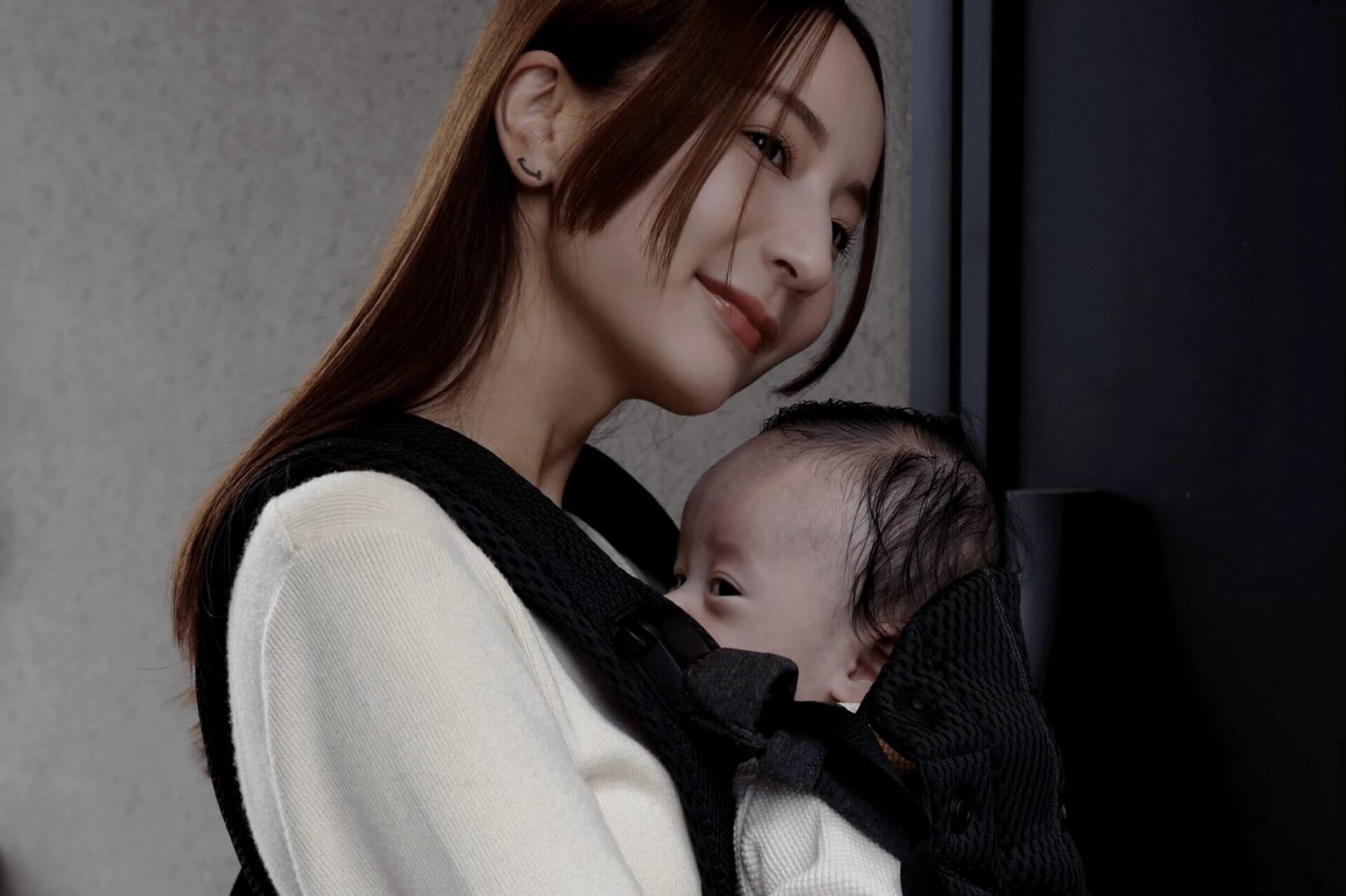 A woman with long hair smiles gently while holding a baby in a black carrier. The background is a neutral wall.
