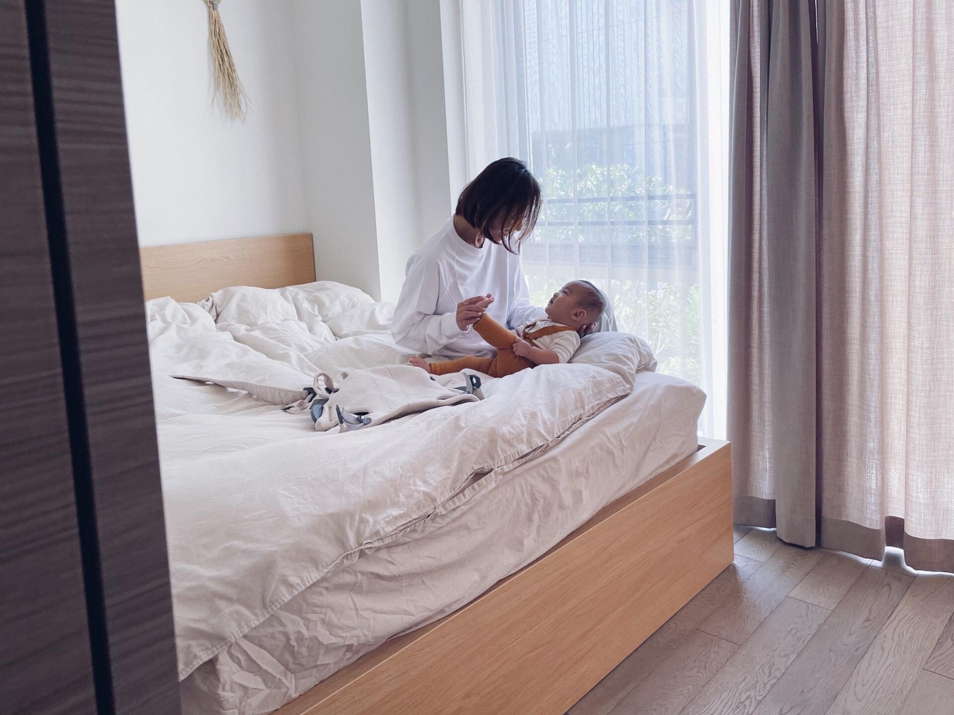 A woman sitting on a bed with a child, both wrapped in a white blanket, in a softly lit room with curtains and a window.