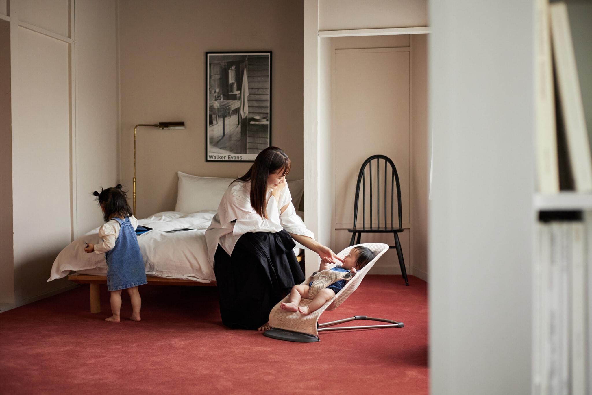 A woman tends to a baby in a bouncer while a toddler stands nearby in a cozy bedroom with a bed, chair, and framed poster on the wall.