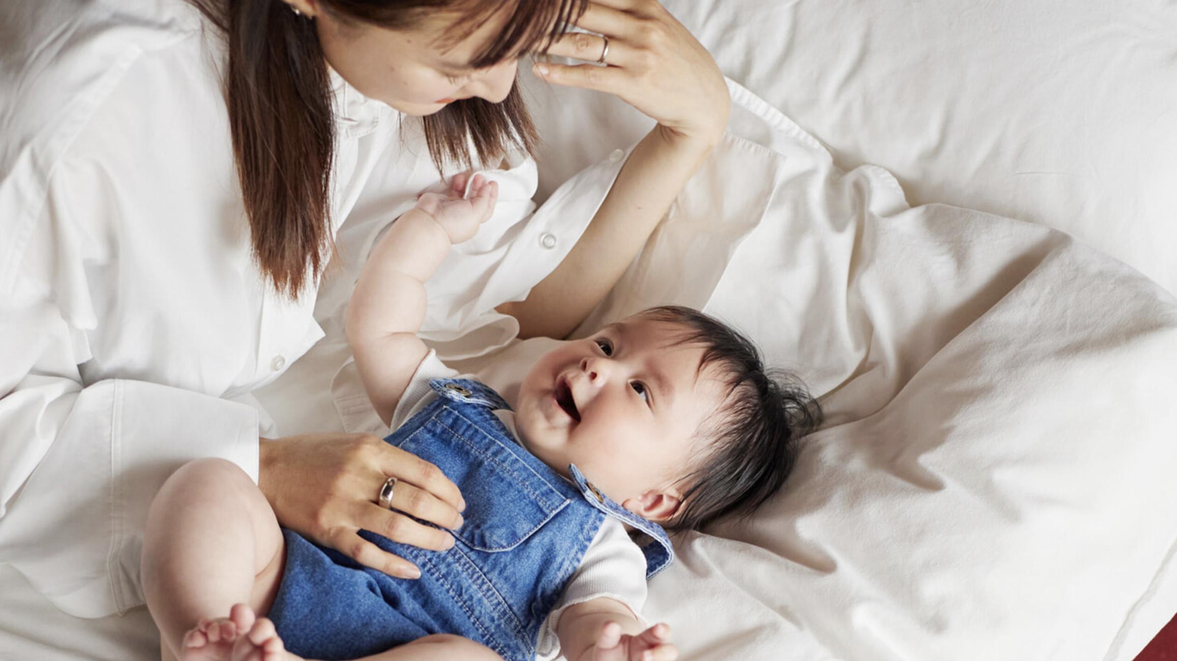A woman in a white shirt smiles at a baby wearing denim overalls, lying on a white bed, both appearing happy and engaged.