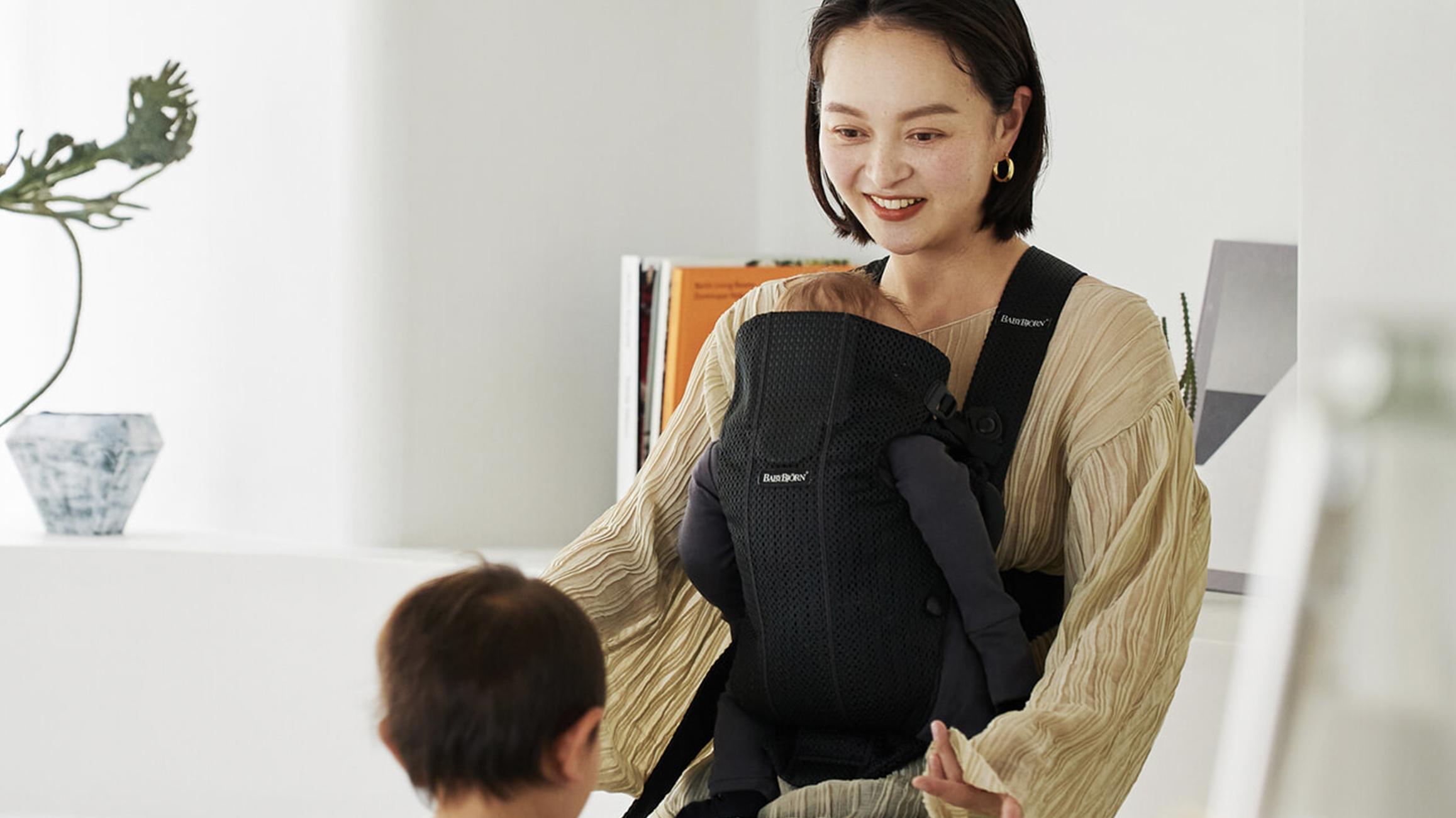 A woman smiles while holding a baby in a black carrier. Another child stands nearby in a bright, minimalistic room.