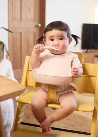 赤ちゃんが黄色いハイチェアに座り、ピンクのスタイをつけてスプーンを持っている。背景に木製のドアとテレビがある。
