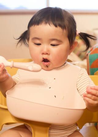 食事用の椅子に座り、ピンクのビブをつけた幼児がスプーンを持って食事をしている様子。髪はツインテール。