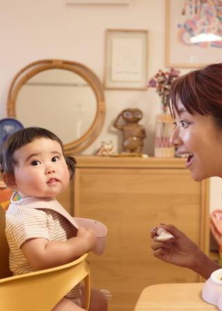女性と赤ちゃんが食卓で向かい合っている。赤ちゃんは椅子に座り、女性は笑顔で話しかけている。背景に鏡と装飾品。