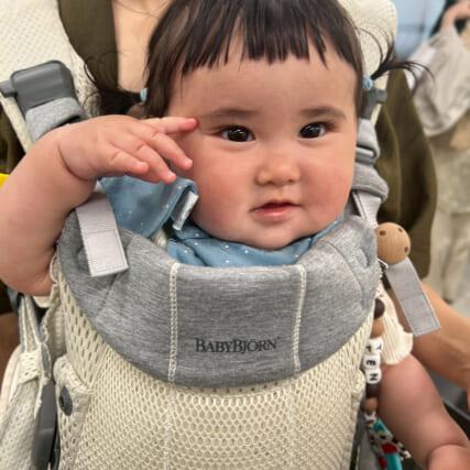 赤ちゃんがベビービョルンのキャリアに乗っている。青いスタイをつけ、周りには大人がいる。赤ちゃんはカメラを見ている。
