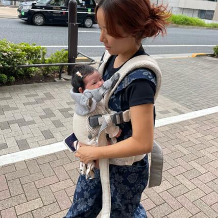 女性が赤ちゃんを抱っこひもで抱え、歩道を歩いている。背景にタクシーが見える。