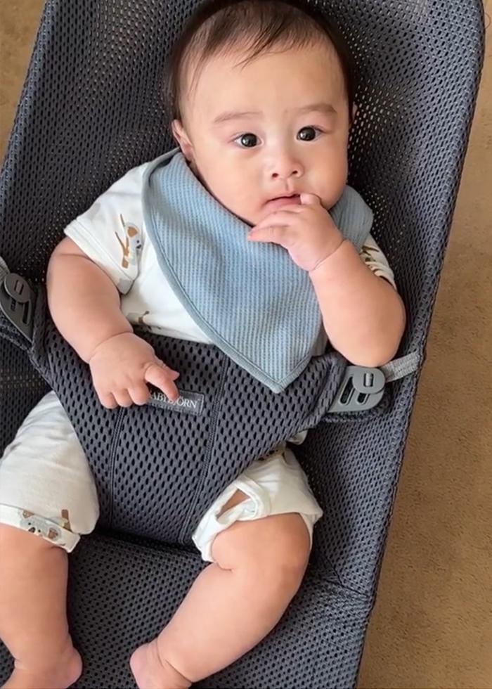 A baby wearing a blue bib and white outfit sits in a gray mesh baby bouncer, with one hand near their mouth, looking curious.