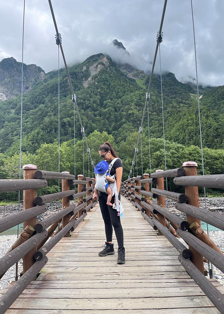 A woman carrying a baby stands on a wooden suspension bridge, surrounded by lush green mountains under a cloudy sky.
