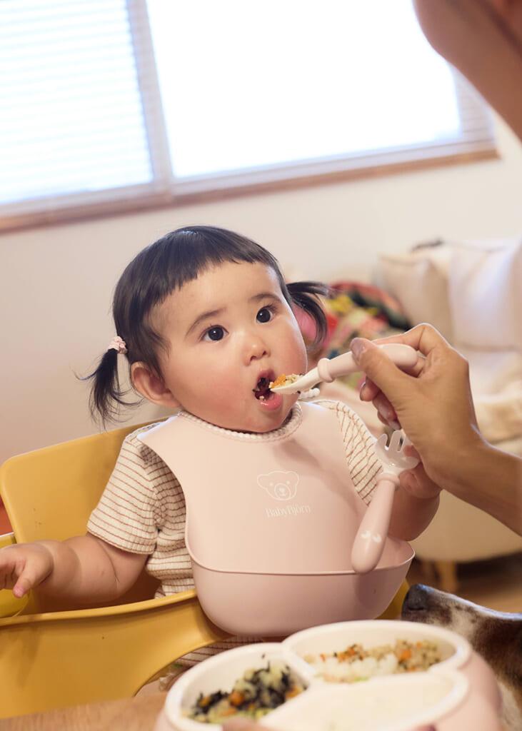 幼児がピンクのビブをつけて椅子に座り、大人がスプーンで食事を与えている。背景に窓がある。
