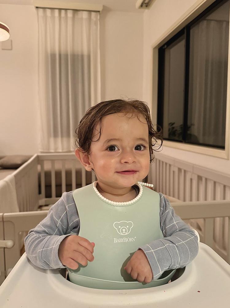 A smiling toddler with wet hair sits in a high chair wearing a light blue bib. The room is softly lit with a crib and window in the background.