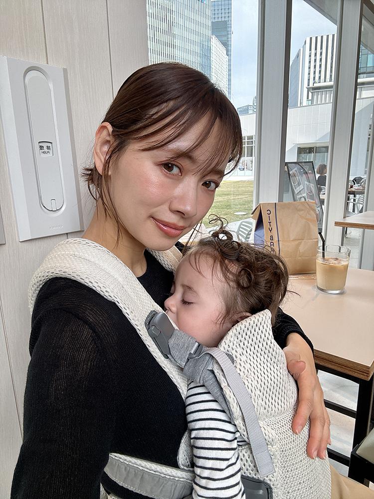 A woman smiles while holding a sleeping baby in a carrier. They are indoors near a window with city buildings visible outside.