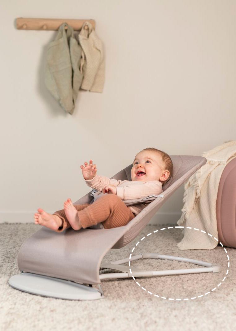 A baby smiling and reclining in a gray baby bouncer on a carpeted floor, with clothes hanging on a wall hook in the background.