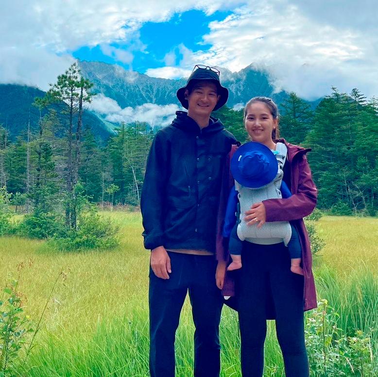 A couple stands in a grassy field with mountains and clouds in the background, holding a baby in a carrier.