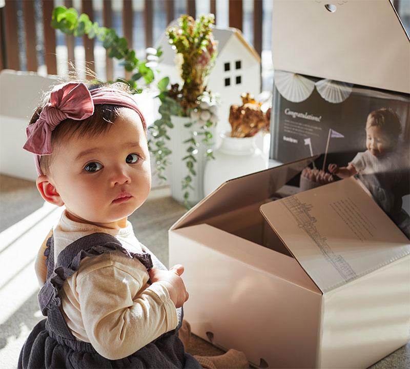 A baby with a pink bow headband sits beside an open box, looking curiously. There's a small white house and plants in the background.