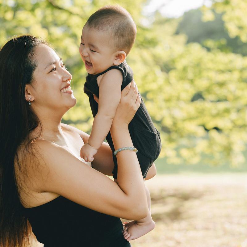 A woman joyfully holds a laughing baby outdoors, surrounded by lush green trees on a sunny day.