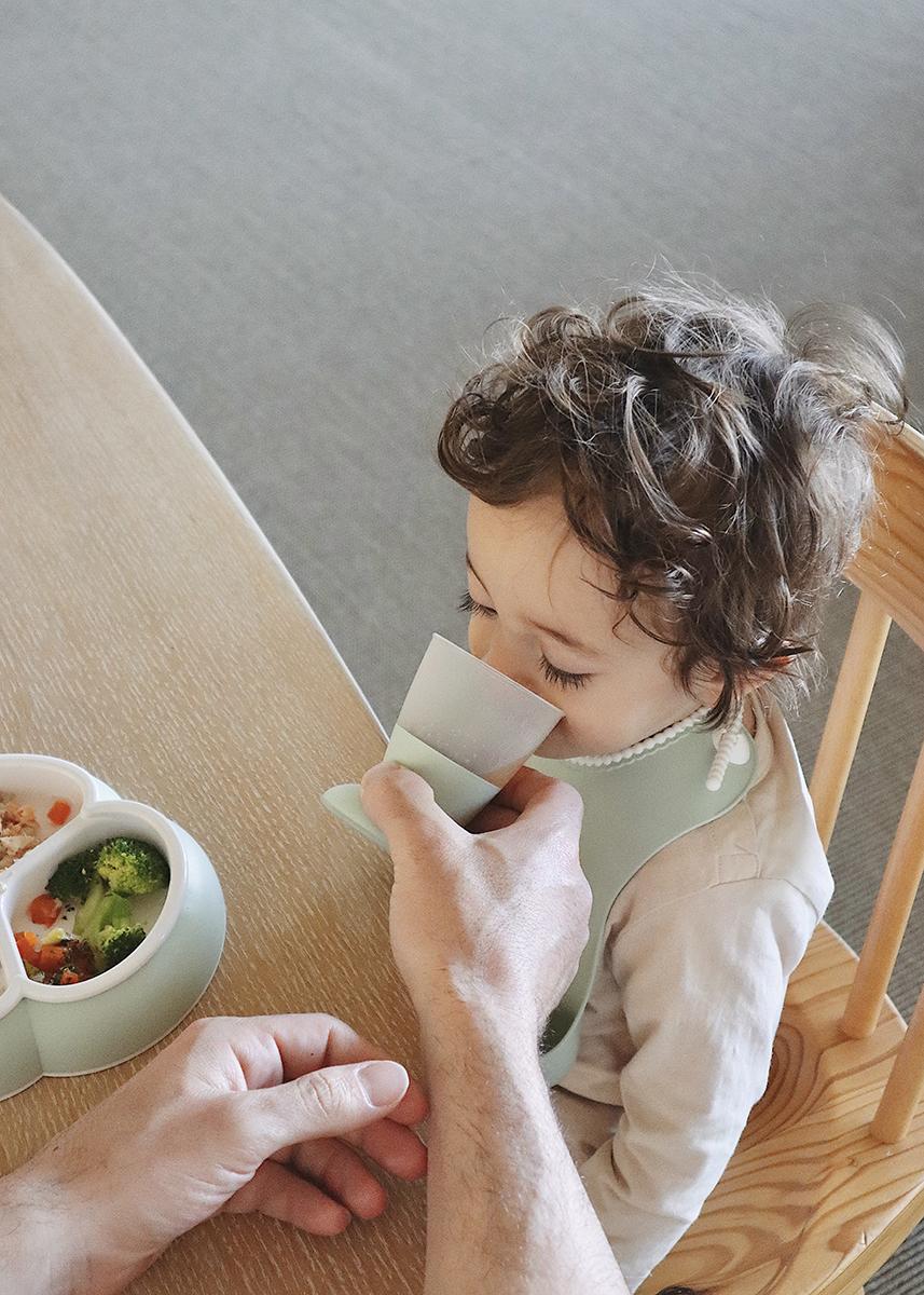 A toddler with curly hair drinks from a cup held by an adult at a wooden table with a plate of food, including broccoli, nearby.