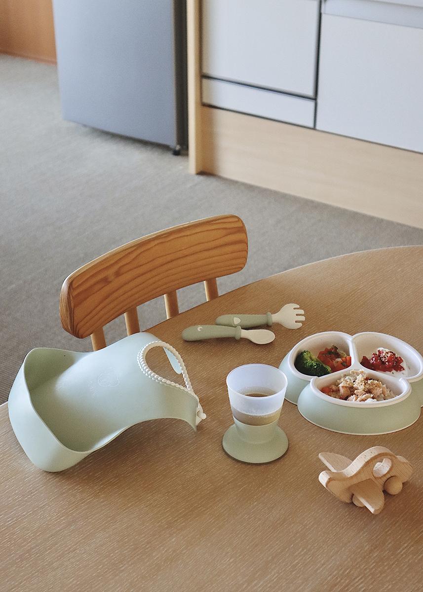 Child's dining setup with a silicone bib, divided plate with food, utensils, a cup, and a wooden toy on a wooden table.