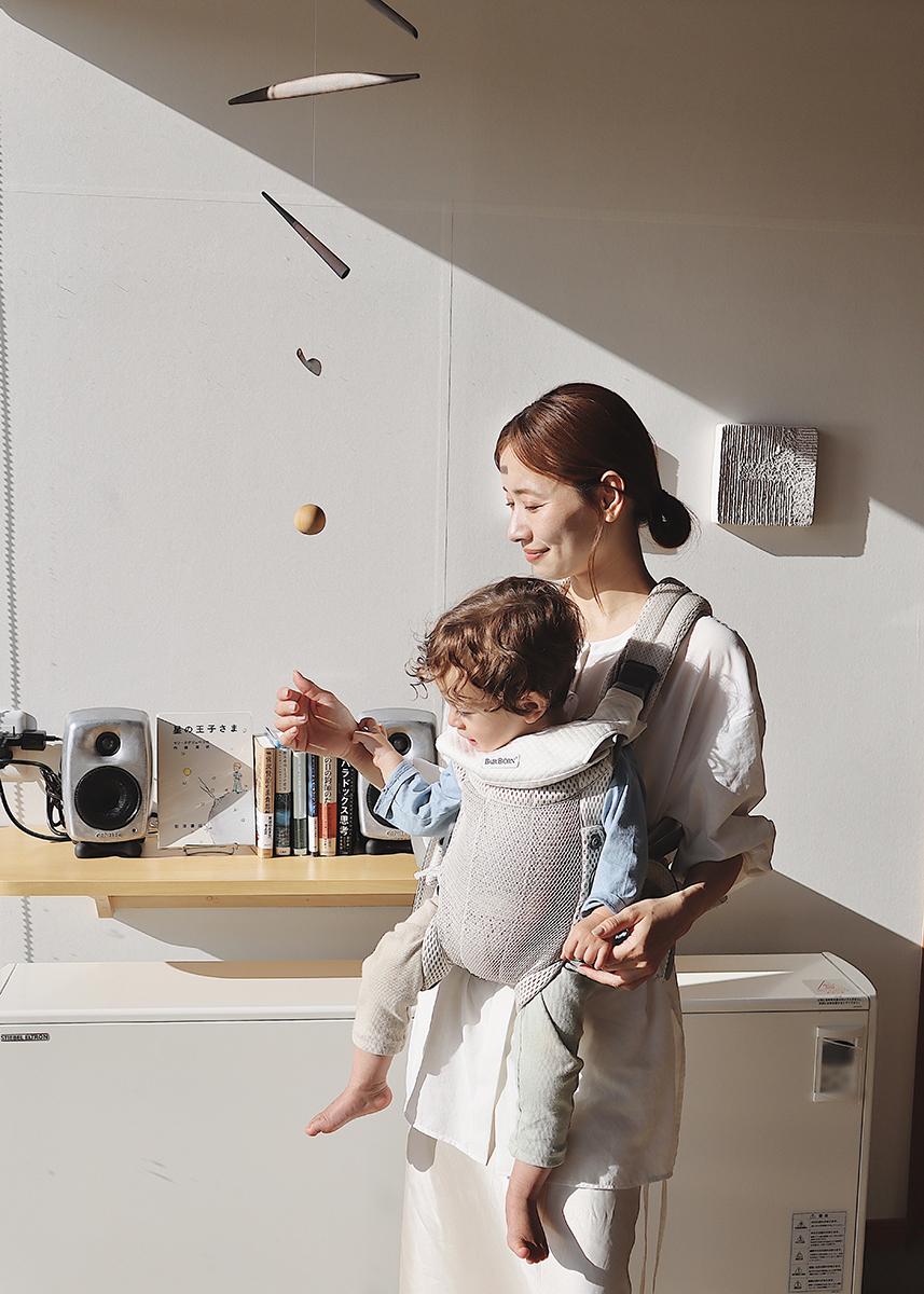 A woman stands in a sunlit room, holding a child in a baby carrier. They are near a shelf with books and speakers, reaching towards a mobile.