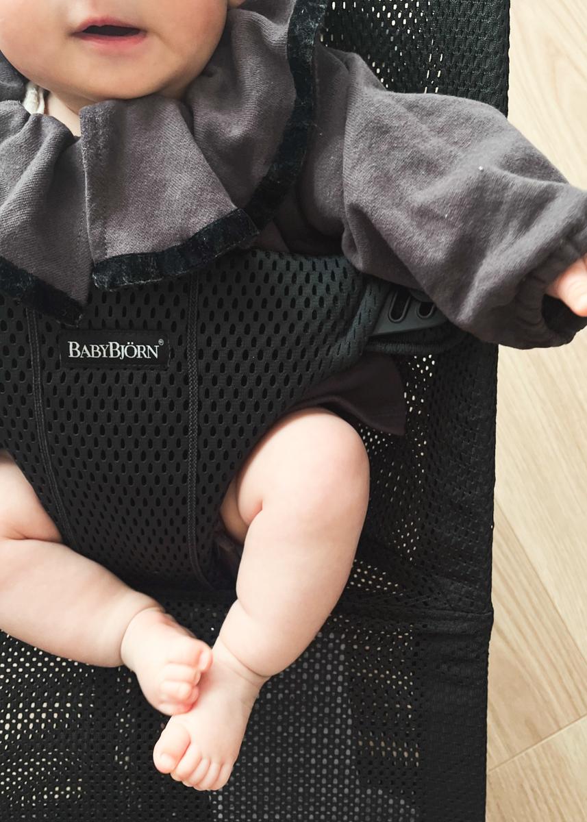 A baby in a dark outfit sits in a black BabyBjörn bouncer, with only the lower half of the face and legs visible.
