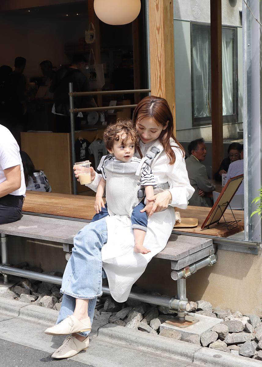 Woman sitting on a bench outside a café, holding a toddler in a carrier. Both are smiling and enjoying drinks.