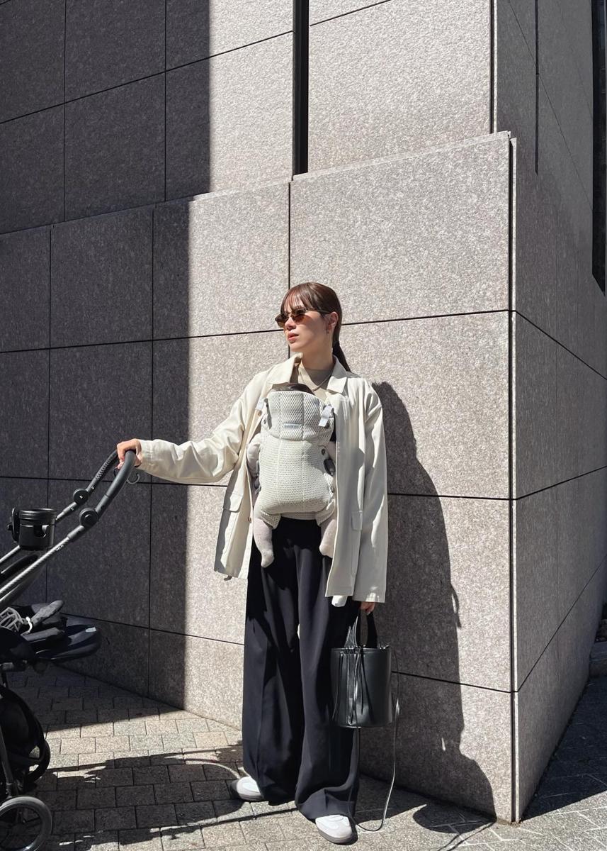 Person wearing sunglasses, holding a stroller, with a baby in a carrier, stands against a modern stone building in sunlight.