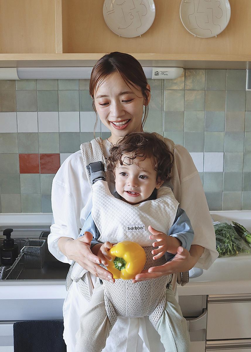 Smiling woman holding a toddler in a baby carrier, both holding a yellow bell pepper in a kitchen with tiled backsplash.