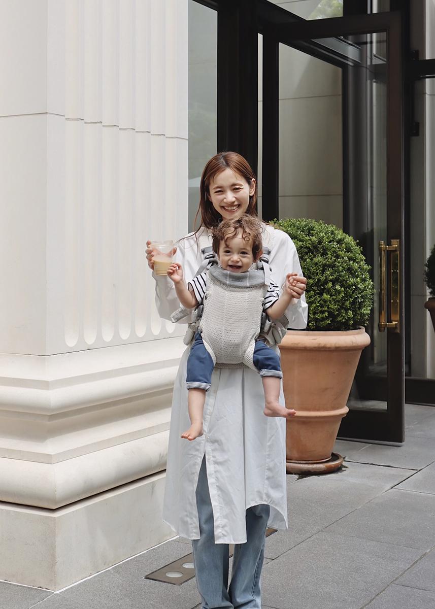 A woman smiling while holding hands with a baby in a front carrier, standing outside a building with large potted plants.