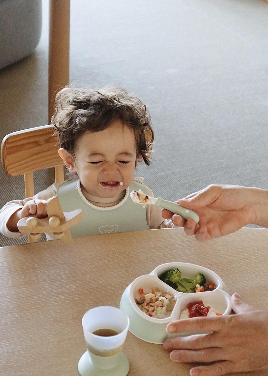 A toddler in a high chair makes a funny face while being fed. A divided plate with food and a cup are on the table.