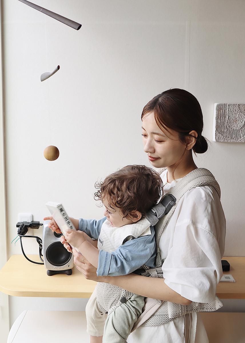 A woman holds a baby in a carrier, both looking at a small white object. A mobile hangs nearby, and a wooden shelf is in the background.