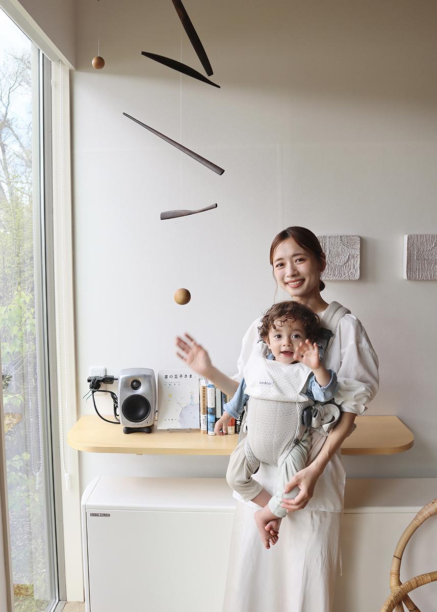 A woman smiling and holding a child in a carrier, standing by a window with a small shelf, books, and a speaker.