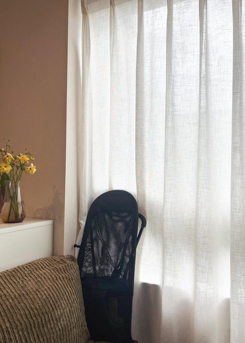 A cozy room with sheer curtains letting in light, a textured sofa, a vase of yellow flowers on a white cabinet, and a black mesh bouncer.