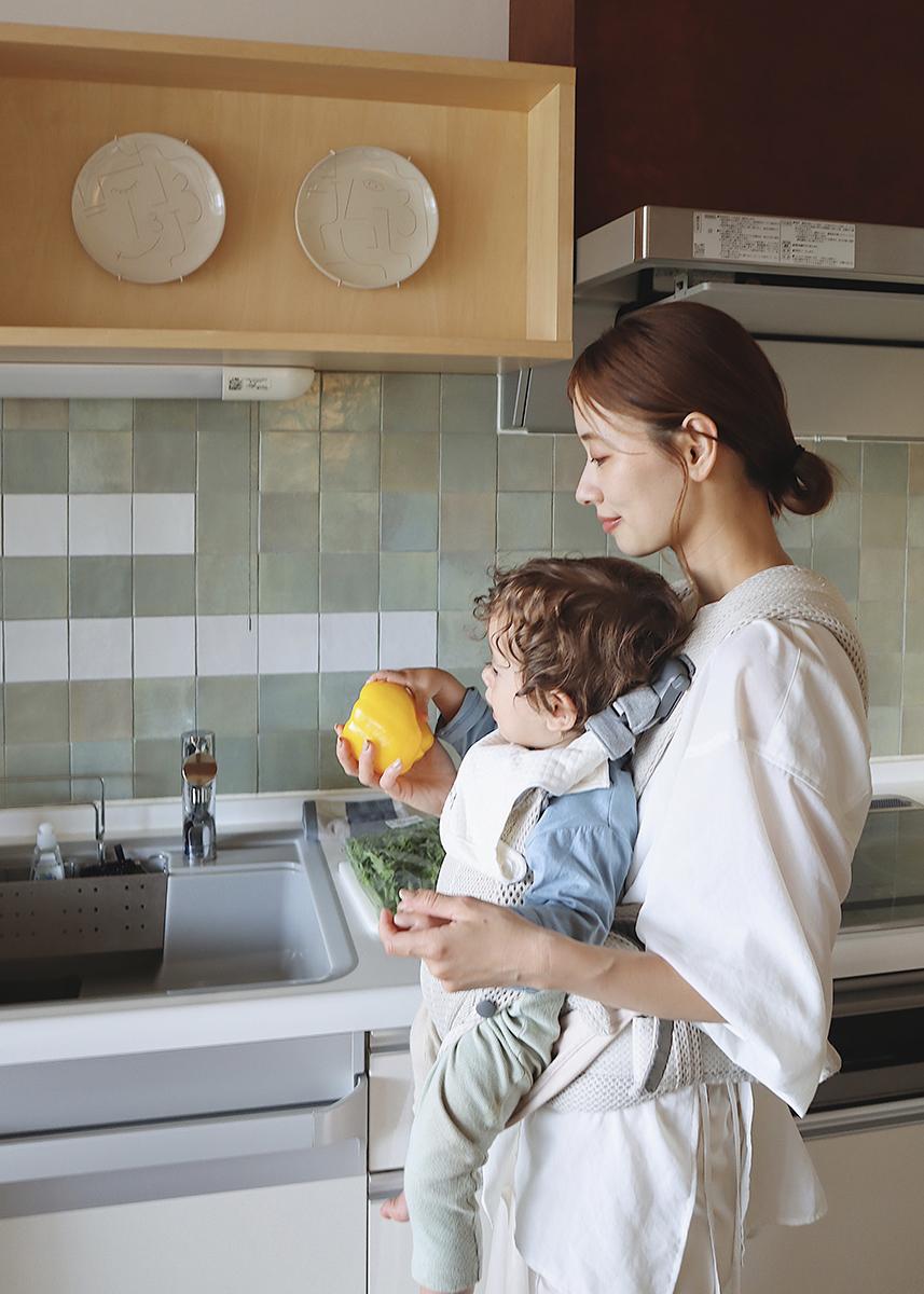 A woman holding a baby in a kitchen, examining a yellow bell pepper, with green vegetables on the counter and tiled backsplash.