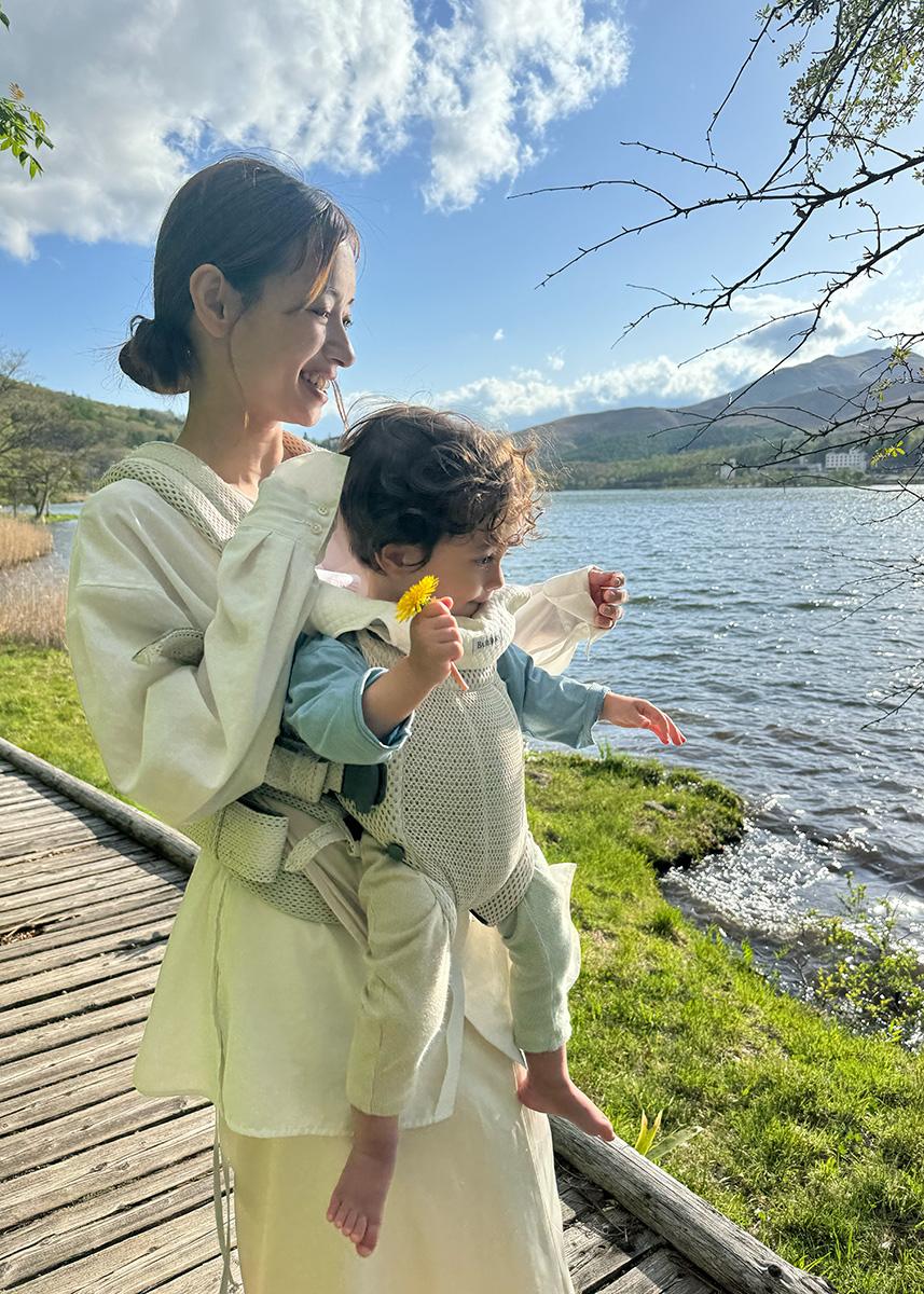 A woman smiles while carrying a child in a baby carrier by a lake. The child holds a yellow flower, and the sun shines brightly.
