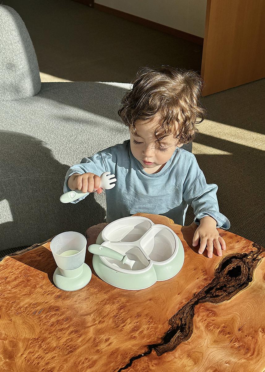 Young child in a blue shirt sits at a wooden table with a divided plate, cup, and fork, appearing to eat or play with the utensils.